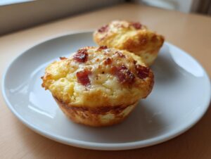 Two freshly baked Pizza Muffins topped with melted cheese and crispy bacon pieces, served on a white plate.
