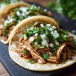 Close-up of two soft corn tortilla chicken tacos filled with shredded chicken, topped with diced white onion and fresh cilantro.