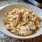 A close-up of shredded slow cooker garlic parmesan chicken coated in a creamy, herby sauce, served in a white bowl.