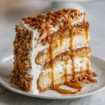 A tall slice of caramel crunch cake layered with cream, topped with crunchy nuts and drizzled heavily with caramel sauce.