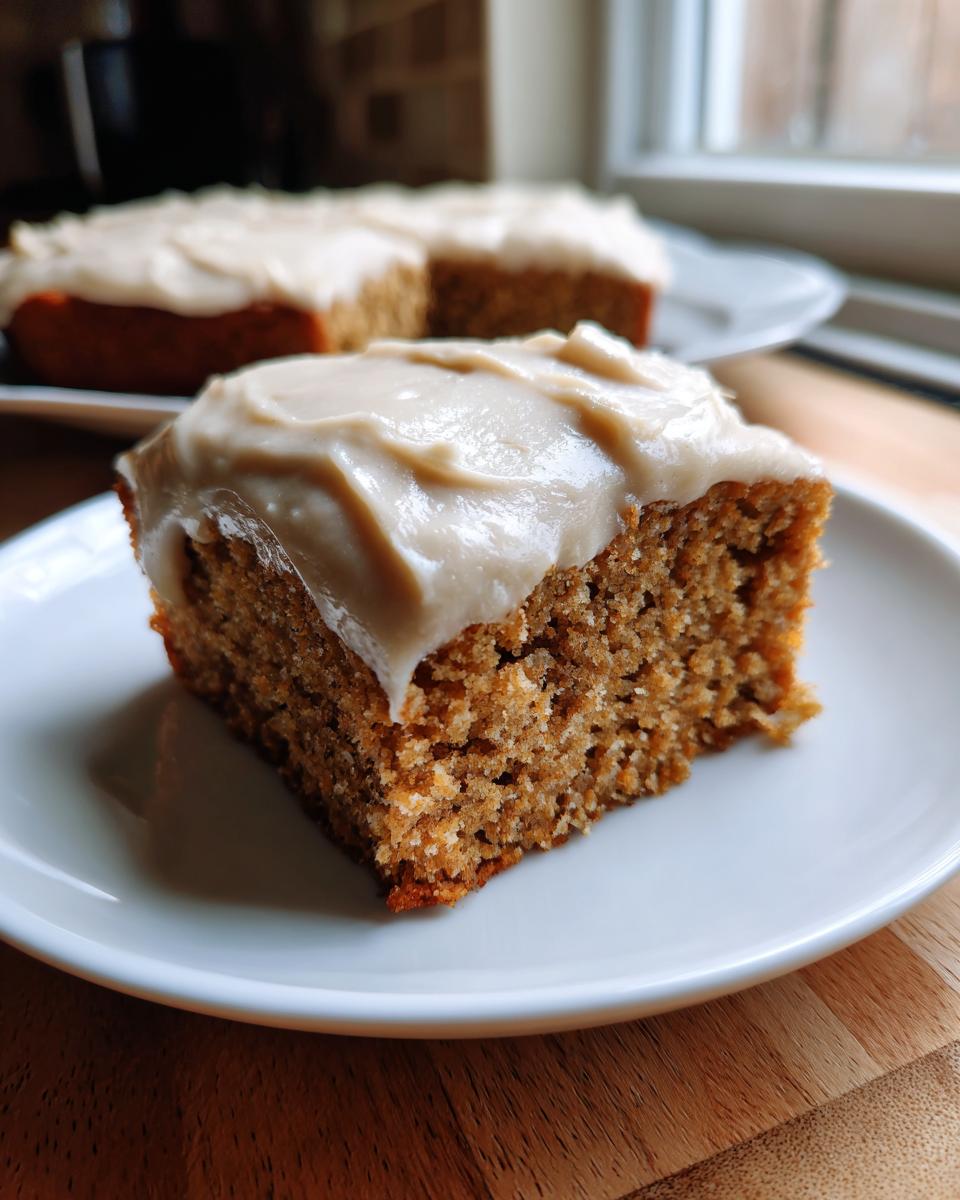 A close-up of a moist slice of applesauce cake topped with thick, creamy frosting, with the rest of the cake blurred in the background.