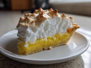 A close-up of a thick slice of lemon meringue pie showing bright yellow filling and toasted meringue peaks.