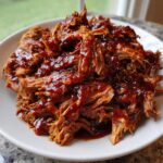 A close-up of shredded, tender Slow Cooker Pulled Pork heavily coated in glossy barbecue sauce, served on a white plate.