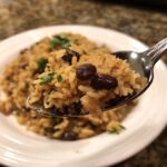 A close-up shot of a spoonful of seasoned rice mixed with dark black beans and fresh cilantro, lifted above a plate of black beans and rice.