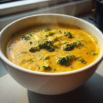 Close-up of a steaming white bowl filled with rich, creamy, orange-yellow broccoli cheddar soup with visible florets.