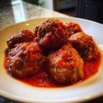 Five tender Italian meatballs smothered in rich, red marinara sauce served on a white plate.