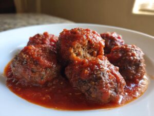 Close-up of several tender Italian meatballs covered in thick, bright red tomato sauce on a white plate.