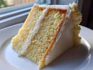 A close-up slice of moist vanilla cake recipe layered with white frosting, sitting on a white plate.