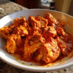 Close-up of rich, orange-red butter chicken pieces coated in creamy sauce, served in a white bowl.