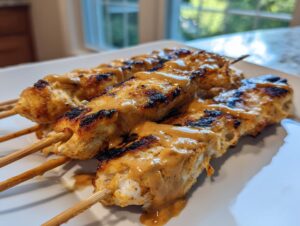Three skewers of grilled chicken satay drizzled generously with creamy peanut sauce on a white plate.