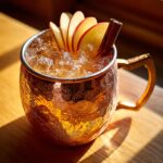 A close-up of a chilled apple cider moscow mule cocktail garnished with apple slices and a cinnamon stick in a hammered copper mug.