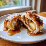 A warm apple dumpling cut in half, showing the spiced apple filling and drizzled generously with caramel sauce.