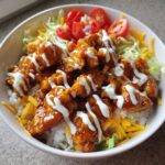 A close-up of a buffalo chicken bowl featuring saucy chicken pieces over rice, topped with ranch, lettuce, tomatoes, and shredded cheese.