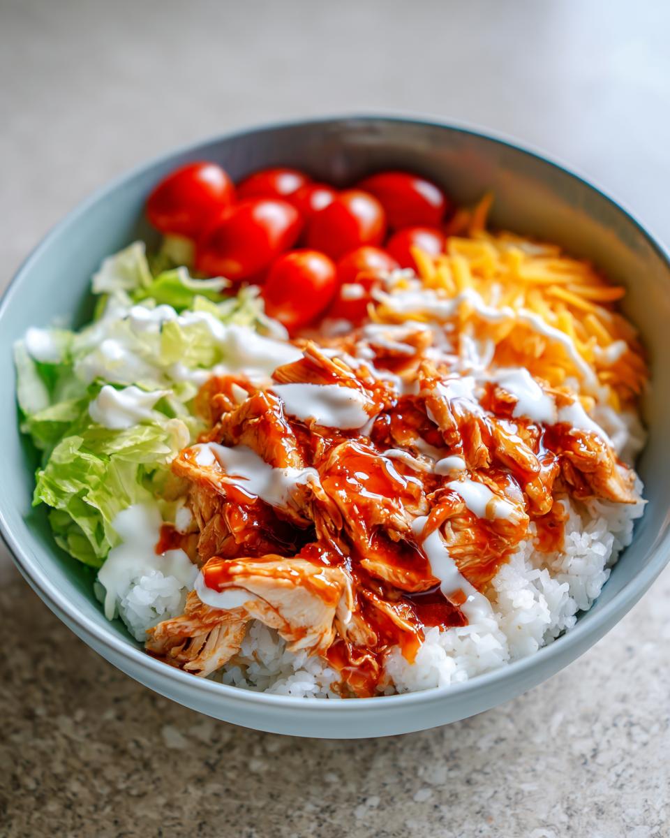 A vibrant buffalo chicken bowl featuring shredded chicken tossed in sauce over white rice, topped with ranch, lettuce, tomatoes, and cheese.