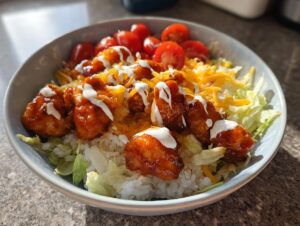 A close-up of a delicious buffalo chicken bowl featuring saucy chicken pieces over rice, shredded lettuce, cheese, and cherry tomatoes.