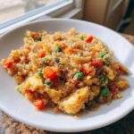 A close-up of a white bowl filled with healthy cauliflower fried rice mixed with scrambled egg, diced carrots, and green peas.