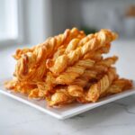 A tall stack of golden brown, flaky, twisted cheese straws recipe on a white rectangular plate.