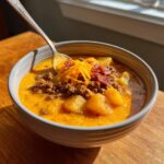 A close-up of a bowl of creamy, orange-hued cheeseburger soup topped with ground beef, shredded cheese, and bacon bits.
