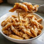 A spoonful of penne noodles covered in melted, stringy cheese being lifted from a bowl of rich french onion pasta.
