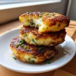 A stack of three golden-brown mashed potato cakes, one broken open to show melted cheese inside, garnished with chives.