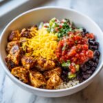 Close-up of a white bowl filled with a colorful chicken burrito bowl featuring seasoned chicken, rice, black beans, shredded cheese, and salsa.