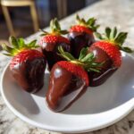 Five fresh, glossy chocolate covered strawberries arranged on a small white plate.