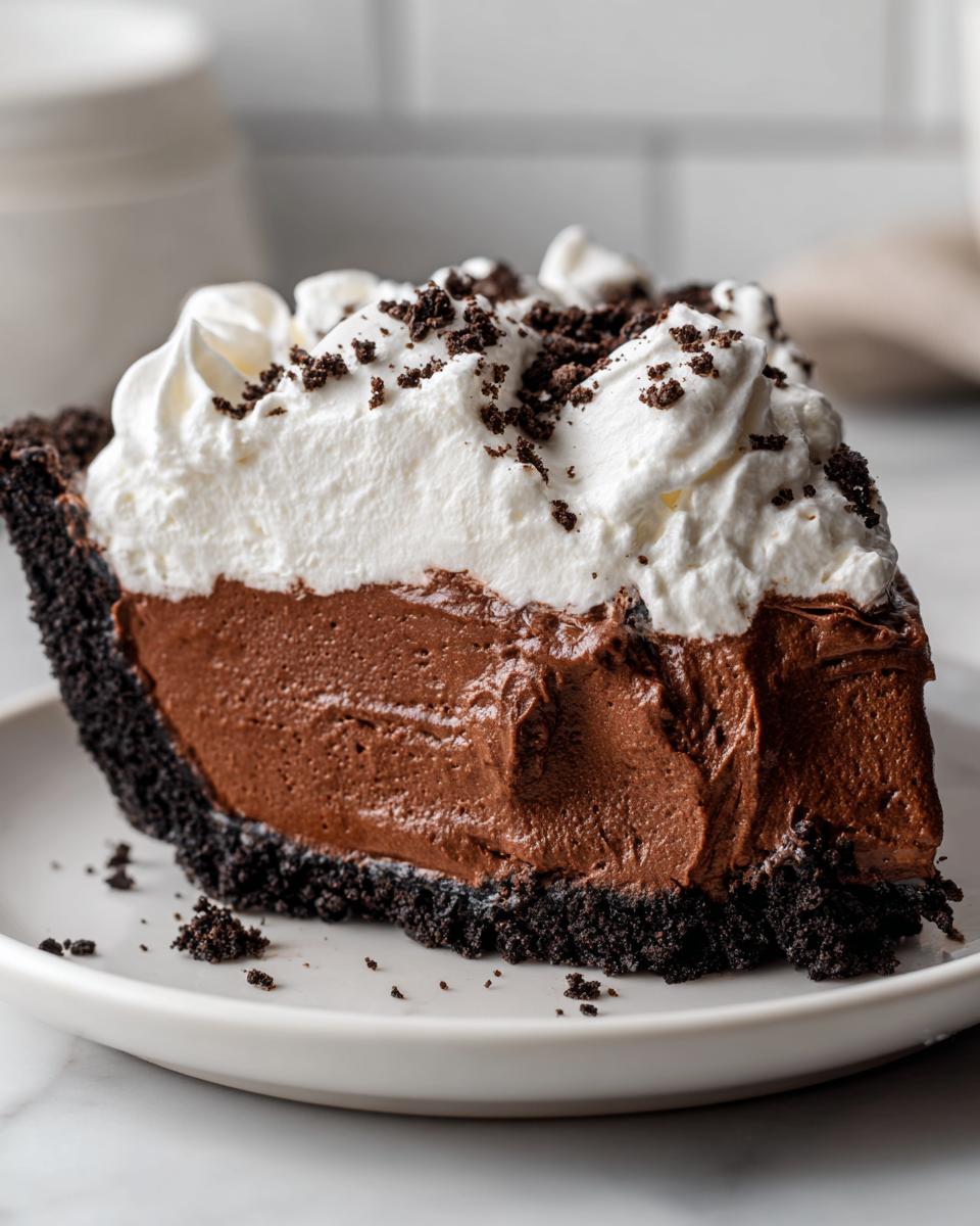 A decadent slice of chocolate mousse pie featuring a dark crust, thick mousse filling, and whipped cream topping sprinkled with crumbs.