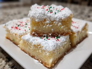 Three squares of Christmas gooey butter cookies stacked and dusted with powdered sugar and red/green sprinkles.