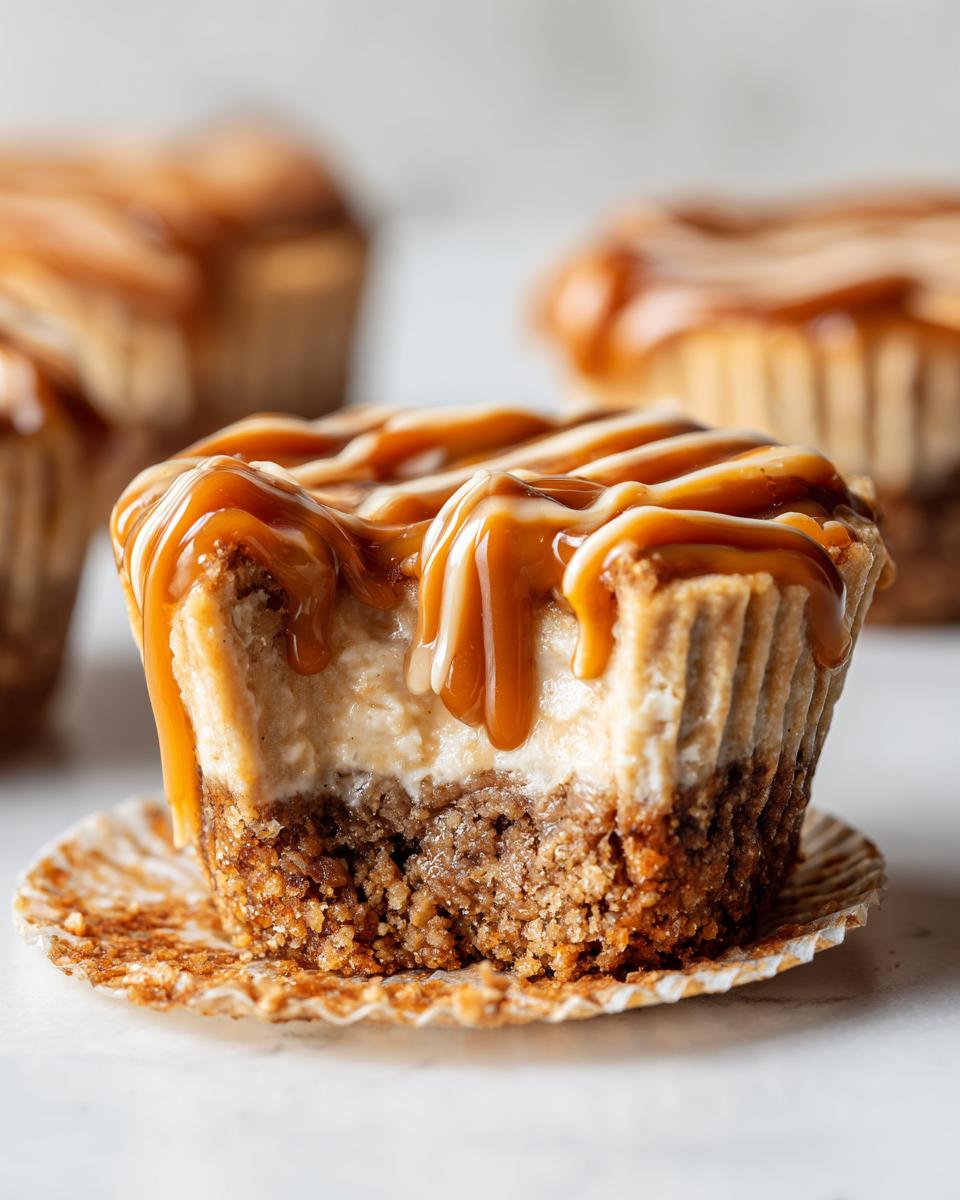 A close-up of a cookie butter cheesecake cup with a bite taken out, showing the crust and creamy filling, topped with a thick caramel drizzle.