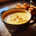 A close-up of creamy, yellow cheese fondue in a dark bowl, topped with croutons and spices, with bread pieces in the background.