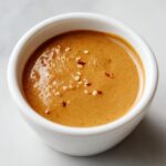 Close-up of creamy peanut sauce garnished with sesame seeds and red pepper flakes in a small white bowl.