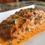 Close-up of a perfectly cooked salmon recipe fillet covered in a rich, creamy, orange-hued sauce with visible herbs.