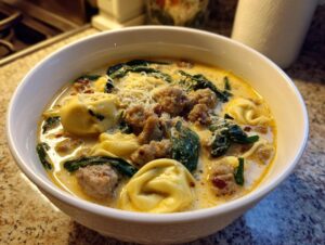 Close-up of a white bowl filled with creamy tortellini soup featuring sausage, spinach, and grated cheese.