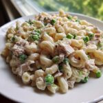 Close-up of a creamy tuna pasta salad featuring elbow macaroni, flaked tuna, and bright green peas served on a white plate.
