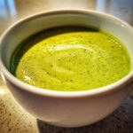 Close-up of vibrant green zucchini soup with a slight swirl on top, served in a white mug.