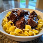 Close-up of tender crockpot beef tips smothered in rich brown gravy served over wide egg noodles in a white bowl.