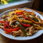A white plate piled high with shredded chicken and colorful strips of sautéed red, yellow, and green peppers, perfect for crockpot chicken fajitas.