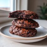 Three fudgy chocolate brownie cookies stacked on a light plate, showing a cracked, shiny top.