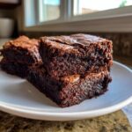 Close-up of three fudgy date brownies stacked on a white plate, showing their rich, dark texture.