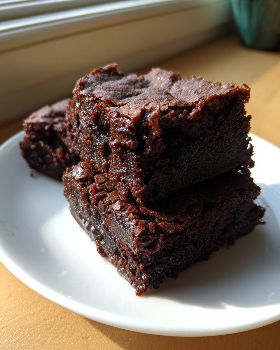 Two stacked, rich, dark, and fudgy date brownies sitting on a small white plate near a window.