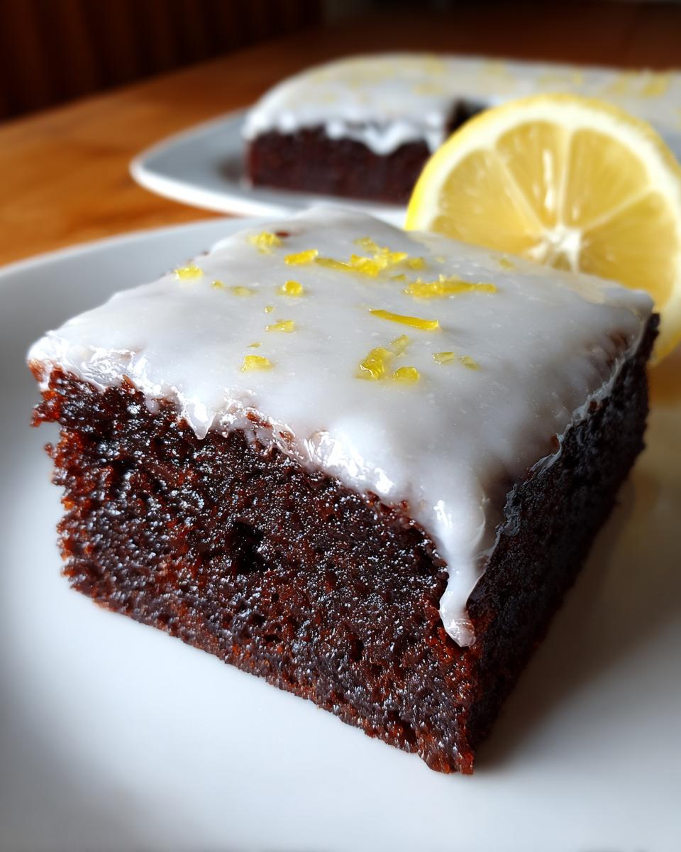 Close-up of a fudgy square of Lemon Brownies topped with white glaze and lemon zest, served next to a lemon slice.