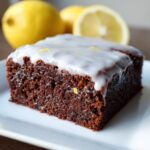 Close-up of a fudgy, dark chocolate square topped with white lemon glaze and zest, with fresh lemons in the background.