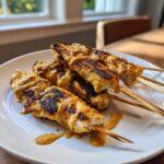 Close-up of grilled chicken satay skewers drizzled generously with peanut sauce on a white plate.