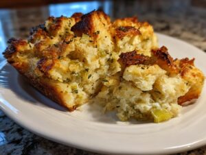 A close-up of a golden brown portion of savory stuffing with visible celery and herbs on a white plate.