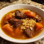 Close-up of a white bowl filled with rich beef barley soup, featuring tender beef chunks, bright orange carrots, and plump barley.