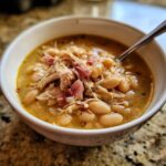 Close-up of a white bowl filled with rich navy bean soup, topped with shredded chicken and ham pieces.