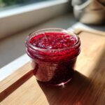 A glass jar filled with vibrant, homemade raspberry jam sitting on a wooden cutting board in bright sunlight.