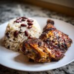Close-up of perfectly seasoned Jamaican jerk chicken served next to a mound of rice and peas on a white plate.