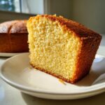 Close-up of a moist, golden slice of lemon olive oil cake on a white plate, with the rest of the cake blurred in the background.