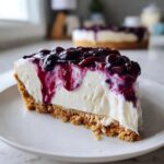 A close-up of a creamy slice of no bake blueberry cheesecake on a white plate, dripping with blueberry topping.